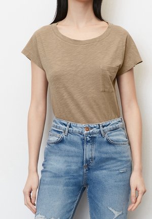 Frau trägt ein beigefarbenes T-Shirt mit kurzen Ärmeln und Tasche, in eine hochtaillierte, hellblaue Jeans mit Used-Look gesteckt, steht vor weißem Hintergrund.