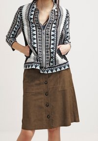 Knopenrok van bruin suède met zijzakken en een middellange snit. Gemanierd met een ritsblouse in blauw en zwart.