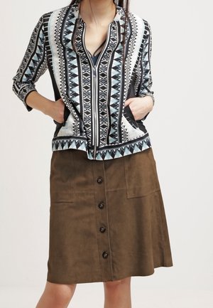 Knopenrok van bruin suède met zijzakken en een middellange snit. Gemanierd met een ritsblouse in blauw en zwart.