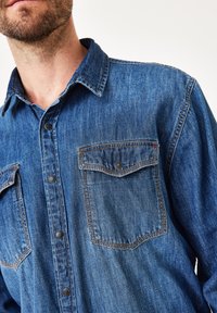 Jeanshemd in Dunkelblau mit klassischem Kragen, zwei Brusttaschen, schwarzen Druckknöpfen und kontrastierenden orangen Nähdetails.