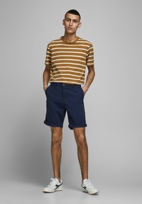 T-shirt rayé marron et crème associé à un short roulé bleu marine. Le modèle porte des baskets blanches avec des accents bleu marine et rouge. Design et coupe simples.