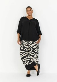 Haut noir ample avec col rabattu ; pantalon zèbre ample en beige et noir ; chaussures noires texturées ; le mannequin se tient droit.
