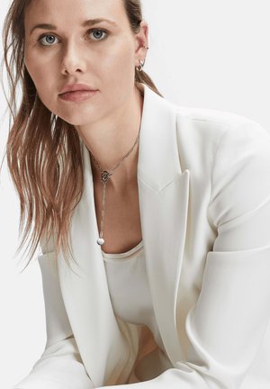 Weißer Blazer mit scharfen Revers, über einem hellen Oberteil getragen. Silberne Kettenhalskette mit Knotendetail und einem hängenden runden Anhänger. Weiche Textur.