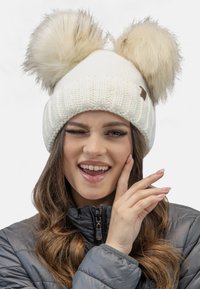 Bonnet en tricot blanc avec deux grands pompons en fausse fourrure crème sur le dessus. Texture côtelée et petite étiquette de marque marron sur le côté.