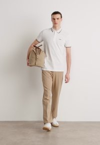 Hvit polo skjorte med svarte detaljer, beige bukser og hvite joggesko. Bærer en lys brun duffelbag med brune stropper, og går mot en ensfarget bakgrunn.