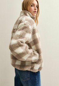 Fleecejacka i beige och vit rutmönster, hög krage, dragkedjestängning, mjukt material, med raglanärmar och sidofickor.