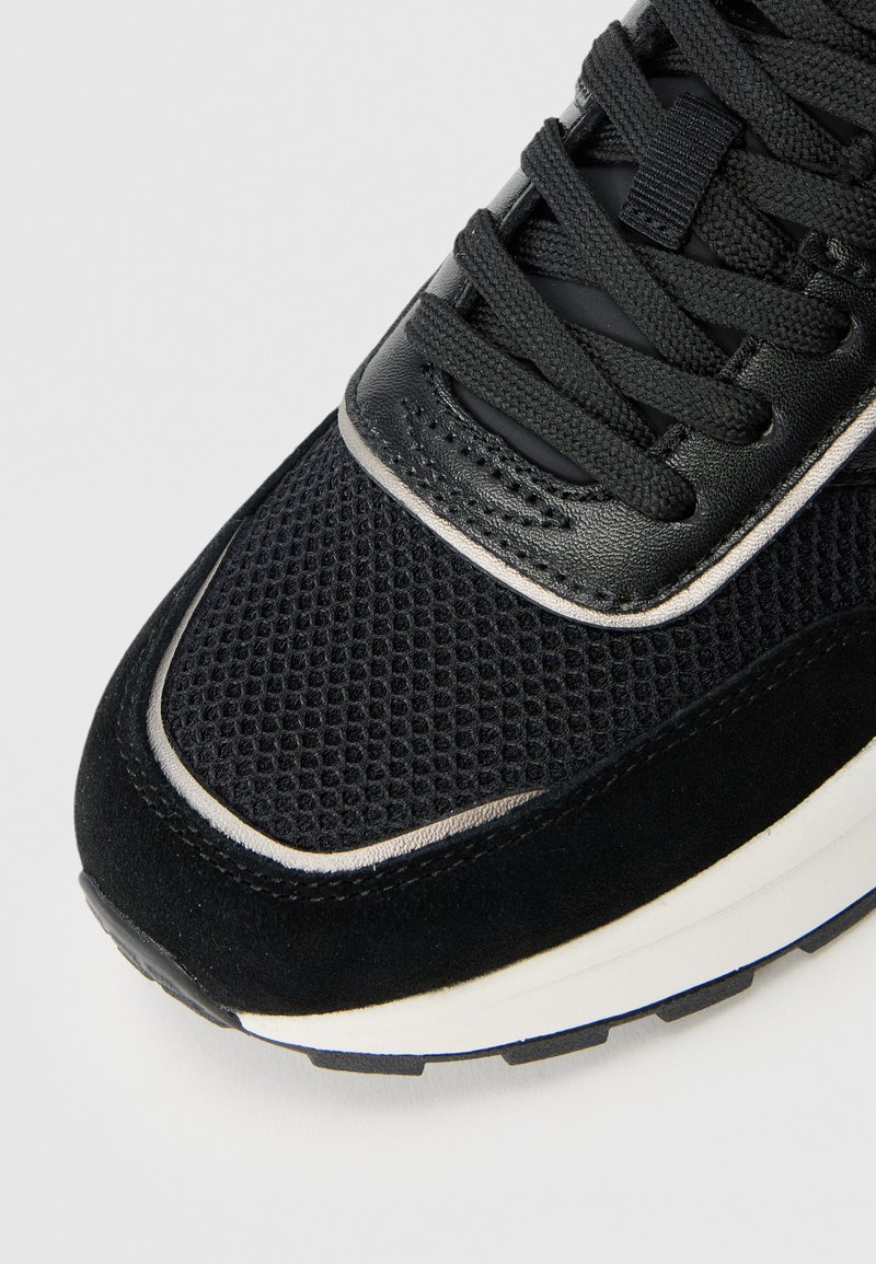 Schwarzer Sportschuh mit einem oberen Teil aus Mesh und Wildlederakzenten, strukturierten Schnürsenkeln und einer dicken weißen Zwischensohle mit Gummisohle.