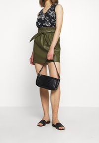Groene faux leren rok met een strikceintuur, gecombineerd met een zwarte tanktop met bloemenprint, en een zwarte croco-embossed handtas. Zwarte slide sandalen.