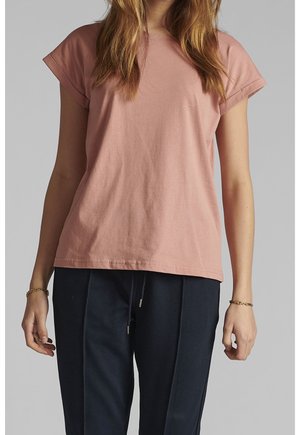 Femme portant une chemise rose à manches courtes ample et un pantalon bleu foncé avec cordon de serrage, debout devant un fond gris uni.