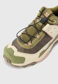 Trail running shoe, olive green and cream upper, mesh panels, textured rubber outsole, lace toggle closure, and pink accents.