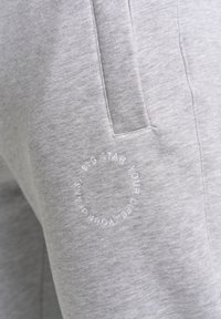 Grå bomulds sweatsbukser med en glat tekstur, der har en sidelomme og et broderet rundt logo med tekst på venstre lår.