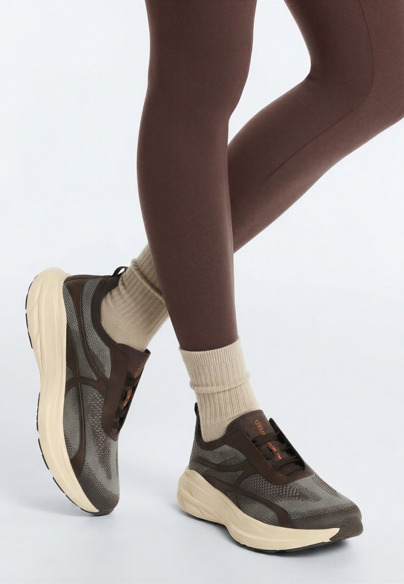 Persoon draagt bruine legging, beige sokken en grijsbruine sportschoenen met dikke crèmekleurige zolen, staand op een lichte achtergrond.