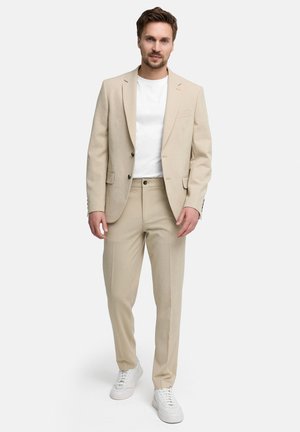 Mand går fremad iført beige jakkesæt, hvid t-shirt og hvide sneakers mod ensfarvet hvid baggrund.