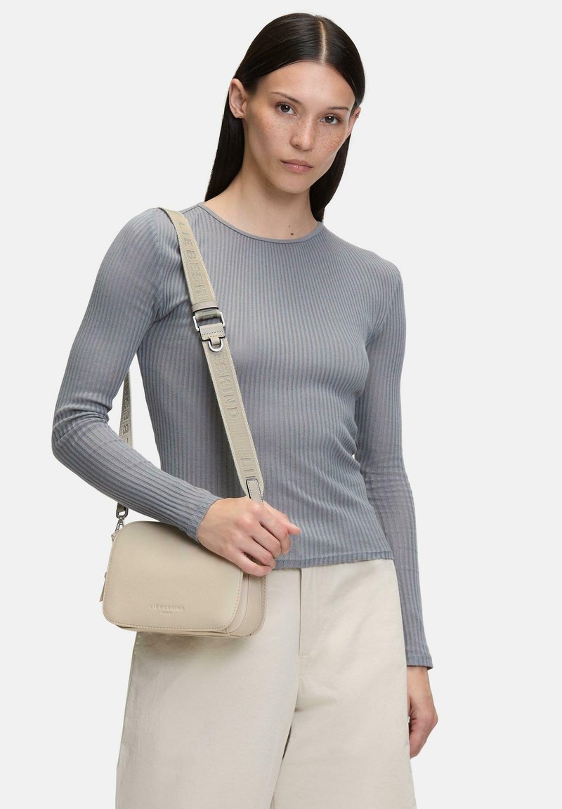 Femme portant un haut gris ajusté à manches longues côtelé et un pantalon beige, portant un petit sac bandoulière beige avec une bandoulière ajustable.
