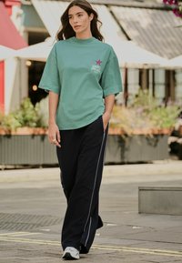 Tricou oversized de culoare verde menta cu un logo în formă de stea, combinat cu pantaloni jogger negri cu dungi albe pe laterale. Modelul merge pe o suprafață exterioară pavata.