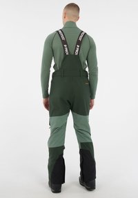 Groene skioverall met verstelbare zwarte banden, met gekleurde panelen in verschillende tinten groen en zwart, en ritsbenen.