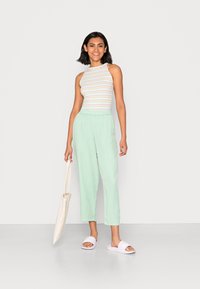 Top de alças às riscas em cores claras, calças largas verdes menta e chinelos brancos. Modelo segura uma mala de mão bege, de pé sobre um fundo simples.
