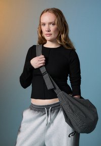 Eine junge Frau mit hellen, welligen Haaren trägt ein schwarzes Langarmoberteil und graue Sweatpants. Sie hält eine große grau-gestreifte Schultertasche.