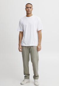White cotton t-shirt with a crew neck, paired with light green pants. Model stands confidently in front of a plain background.