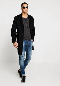 Zwarte wollen jas over een grijze henley, blauwe versleten jeans en zwarte schoenen. Model draagt een zonnebril en staat in een relaxte houding.