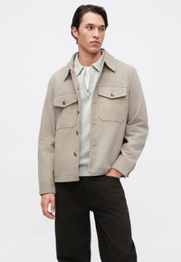 Giacca in suede grigio chiaro con due tasche sul petto e chiusura con bottoni, abbinata a un maglione leggero e pantaloni scuri. Texture liscia e vestibilità su misura.