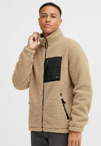 Tan fleece jacket with high collar, front black zip pocket, two side zippers, textured surface, and ribbed cuffs.