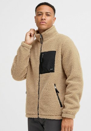 Beige Fleecejacke mit hohem Kragen, schwarzem Reißverschlussfach auf der Vorderseite, zwei seitlichen Reißverschlüssen, strukturiertem Obermaterial und gerippten Bündchen.