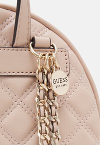 Helle rosa Handtasche aus Kunstleder mit gestepptem Muster, goldfarbene Kettenakzente und einem runden Anhänger mit der Aufschrift "GUESS EST.1981."