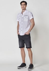 Camisa blanca de manga corta con bolsillos en el pecho abotonados, combinada con pantalones cortos de mezclilla oscuros y enrollados, y zapatillas negras. Diseño simple y funcional.