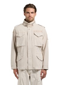 Homme portant une veste de style militaire beige clair avec plusieurs poches à rabat et un col montant, debout devant un fond blanc.