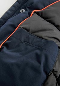 Doudoune marine avec une bordure orange contrastante, présentant une finition texturée et une poche latérale avec un détail de couture visible.