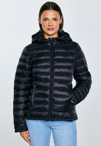 Vrouw die een zwart doorgestikt gewatteerd jack met capuchon en ritssluiting draagt, staand met één hand in haar zak, lichtblauwe jeans, neutrale achtergrond.