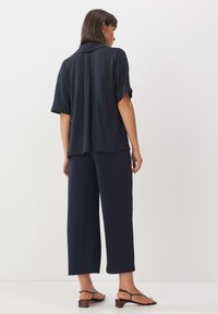 Marineblaue, locker sitzende Bluse mit kurzen Ärmeln und Kragen, kombiniert mit hochtaillierten, weiten Hosen, getragen mit flachen, geschlossenen Sandalen.