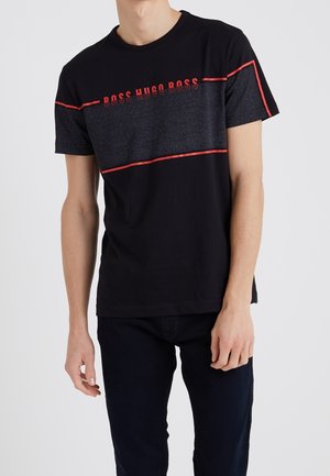 Mężczyzna w czarnej koszulce z krótkim rękawem w czerwone poziome paski, z napisem "BOSS HUGO BOSS" na piersi, w połączeniu z ciemnymi spodniami.
