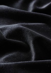 Schwarzer, weicher Samtstoff mit einer glatten Textur, der sanfte Falten und Knitter zeigt. Das Licht spiegelt sich subtil von der Oberfläche wider.