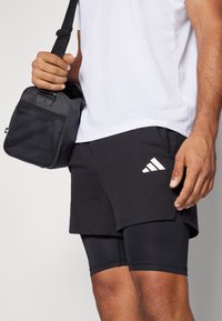 Schwarze Sportshorts mit einer subtilen Textur, die ein weißes Adidas-Logo aufweisen. In einer Hand wird eine passende schwarze Tasche mit Mesh-Details gehalten.