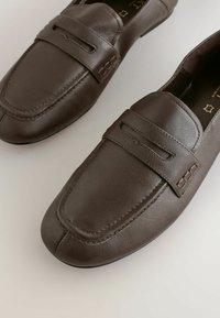 Rudos odinės loafer'iai su siūlėmis ant pirštų, dekoratyvia juostele per pėdą ir glotnia tekstūra, turintys žemą profilį.
