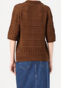 Suéter marrón tejido con un patrón texturizado, mangas cortas y cuello redondo; usado sobre una falda vaquera con bolsillos.