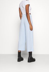 Kvinna klädd i ljusblå vida cropped-byxor, vit cropped t-shirt och svarta ankelskor, stående mot en vit bakgrund.