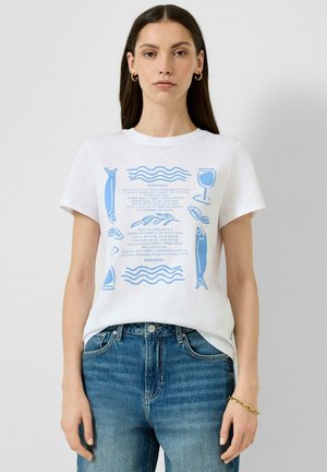 Kobieta w białym T-shircie z niebieską grafiką ryby, kieliszka wina, liści i tekstu, połączonym z niebieskimi dżinsami i złotymi kolczykami w kształcie koła.