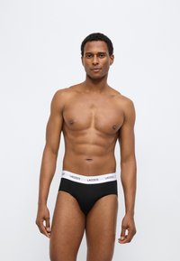 Μαύρα εσώρουχα με λευκή ζώνη μέσης που φέρει το λογότυπο "LACOSTE", παρουσιάζοντας εφαρμοστή σχεδίαση. Το μοντέλο στέκεται σε ουδέτερη στάση.