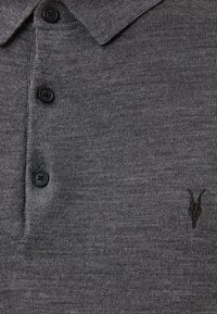 Grå polo skjorte lavet af strikket stof, med en krave, tre sorte knapper og et lille sort broderet motiv på brystet.