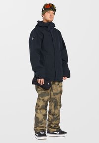 Schwarze Schneejacke mit Kapuze und Reißverschluss, Camouflage-Hose, schwarze Schuhe und eine grüne Mütze mit Goggles. Hält einen schwarzen Handschuh in der Hand.