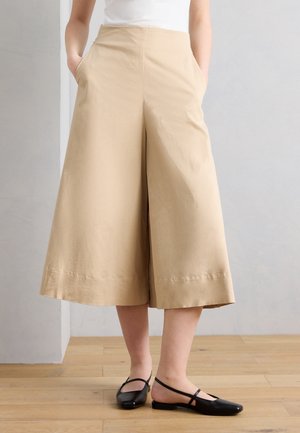 Béžové široké nohavice s krátkou dĺžkou na osobe s rukami v vreckách, doplnené čiernymi balerínami na plochom opätku na drevenej podlahe.