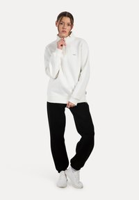 Sudadera blanca de medio cierre con mangas largas, corte relajado y detalle de logo; combinada con pantalones de chándal negros y zapatillas deportivas blancas.