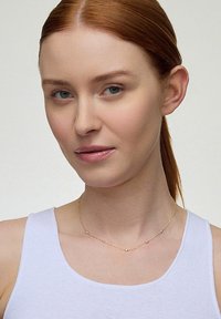 Złoty naszyjnik z małymi okrągłymi koralikami, delikatny łańcuszek, noszony na nagiej skórze nad białym topem na ramiączkach. Gładkie faktury, widoczne drobne detale.