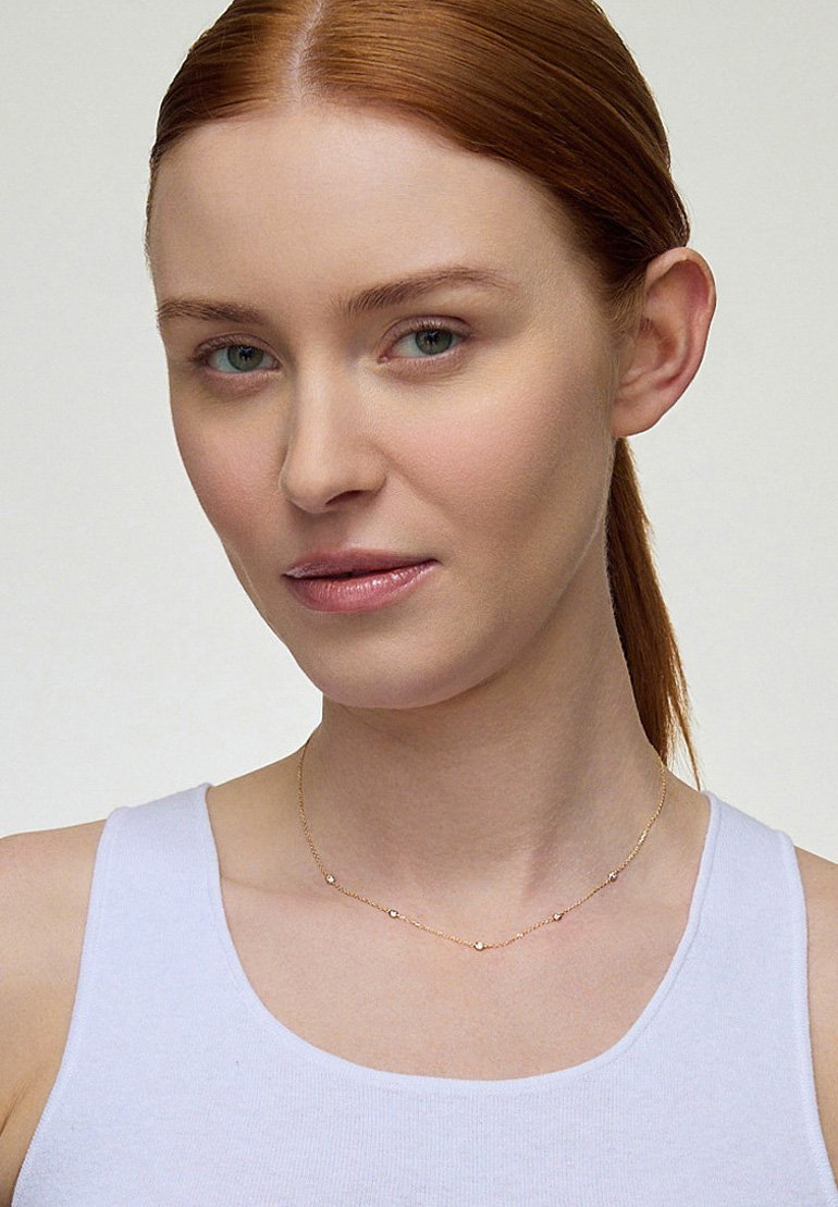 Złoty naszyjnik z małymi okrągłymi koralikami, delikatny łańcuszek, noszony na nagiej skórze nad białym topem na ramiączkach. Gładkie faktury, widoczne drobne detale.