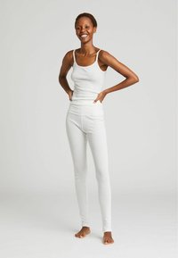 Top a canottiera bianca con coste e spalline sottili, abbinato a leggings aderenti, entrambi realizzati in un tessuto morbido, indossati da una modella che sta in piedi a piedi nudi.