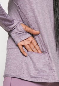 Langærmet, lyse lilla top med lynlås-lomme på siden, lavet af tekstureret stof, med tætsiddende manchetter og kontrastsyede sømme.