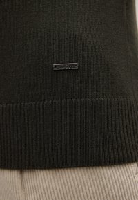 Dunkelgrüner Strickpullover mit geripptem Saum, ausgestattet mit einem silbernen Logo-Tag mit zwei Löchern zur Befestigung, aus weichem Garnmaterial gefertigt.